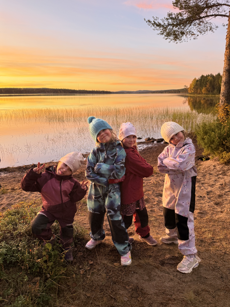 Lapsia poseeraa hymyilleen järven rannalla. Takana auringon lasku maalaa horisonttia oranssiksi.