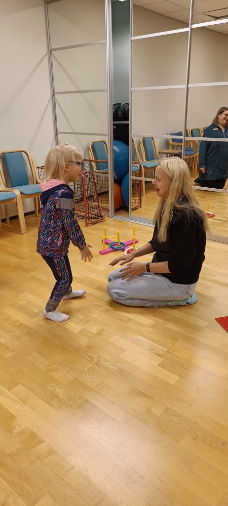 lapsi ja aikuinen liikuntasalissa vastakkain. Aikuinen istuu ja lapsi seisoo. 