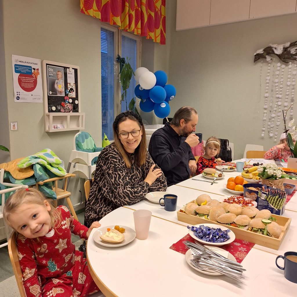 Vanhempia ja pieniä lapsia välipalapöydän ääressä syömässä. Äiti ja lapsi hymyilevät iloisesti kameralle.