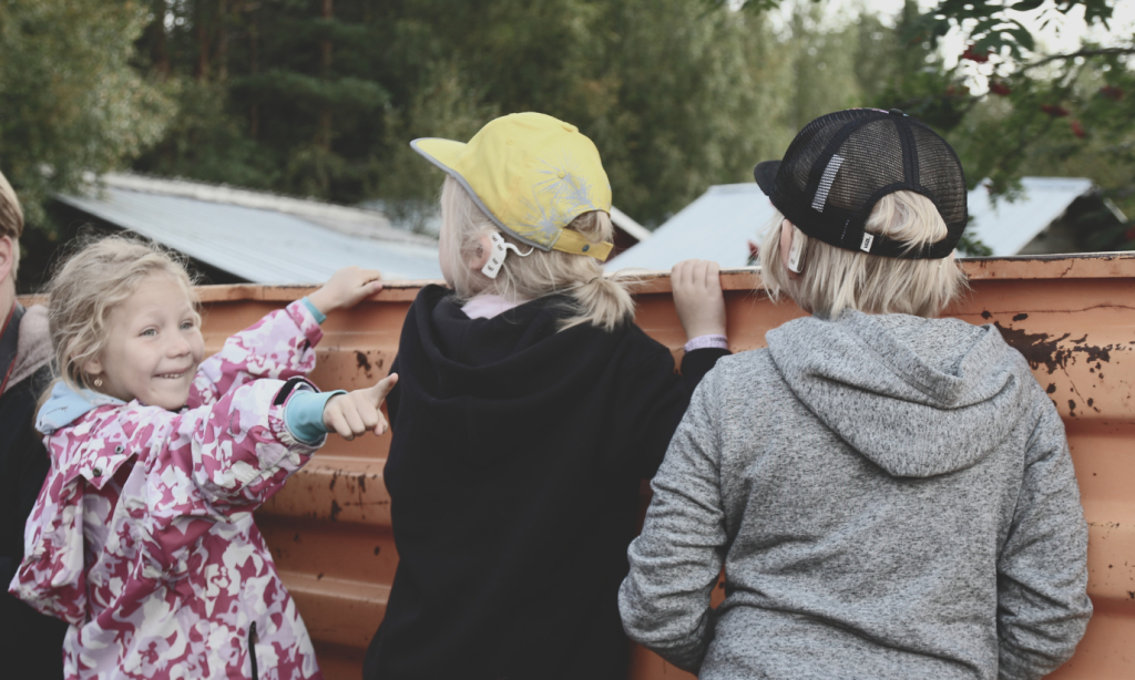 Kolme lasta lavan edessä. Kaksi kääntyneinä lavaa kohti, heillä on sisäkorvaistutteet. Kolmas lapsi katsoo ja osoittaa kuvan ulkopuolelle.