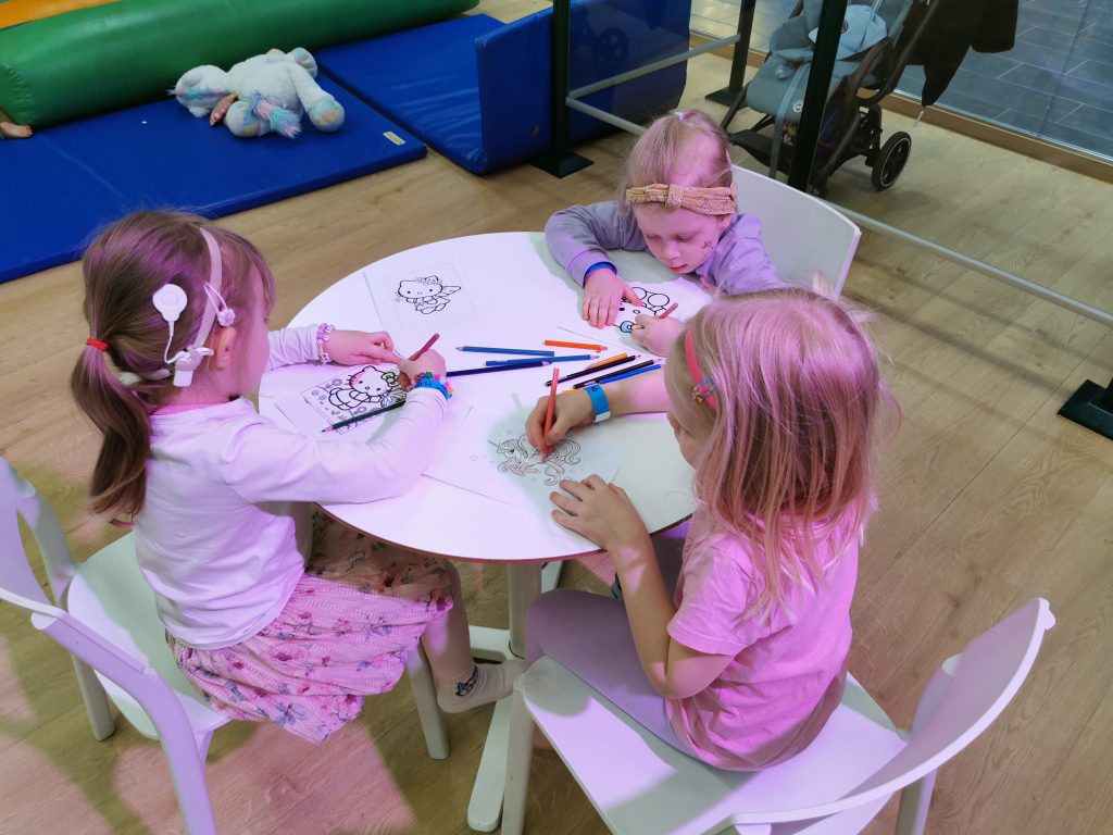 Kolme pientä lasta istuu pöydän ääressä värittämässä värityskuvia. Lapsilla on sisäkorvaistutteet.