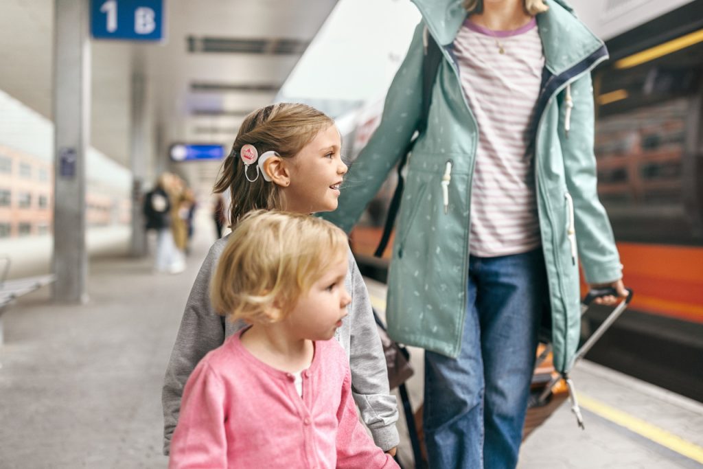 Äiti ja kaksi lasta juna-asemalla, toisella lapsista on sisäkorvaistute.